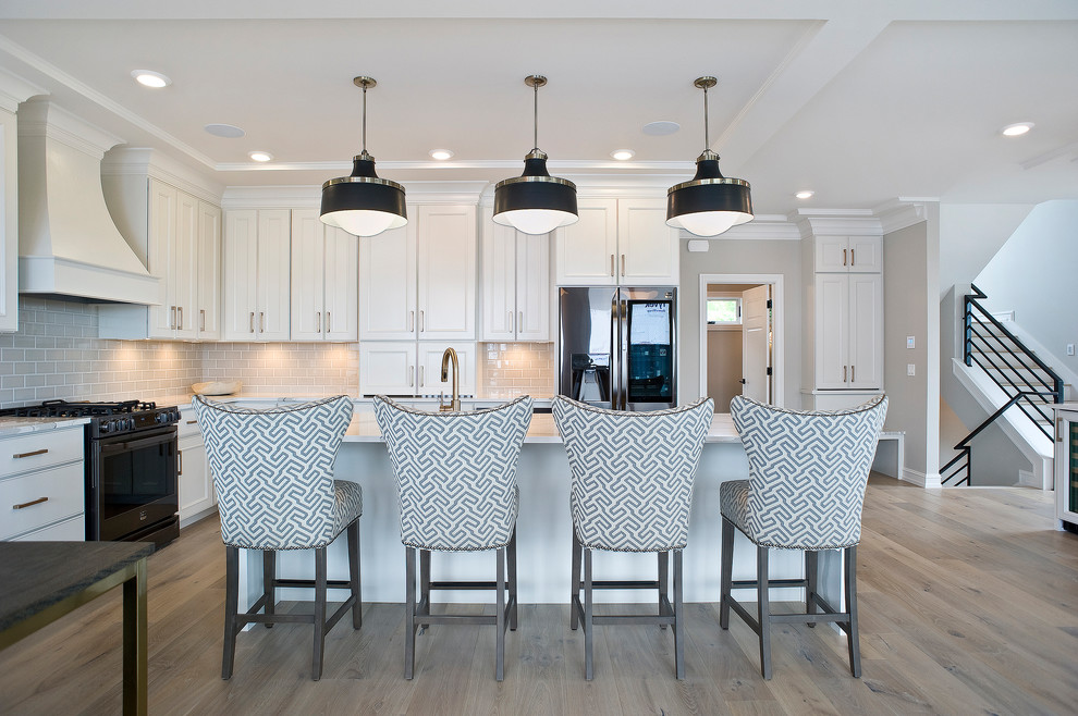 Example of a mid-sized transitional l-shaped light wood floor and brown floor eat-in kitchen design with a farmhouse sink, raised-panel cabinets, white cabinets, marble countertops, gray backsplash, subway tile backsplash, stainless steel appliances and an island