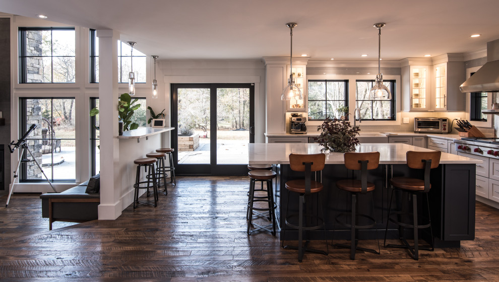 This is an example of a large modern l-shaped eat-in kitchen in New York with a farmhouse sink, shaker cabinets, white cabinets, marble benchtops, white splashback, ceramic splashback, stainless steel appliances, dark hardwood floors, with island, brown floor and white benchtop.