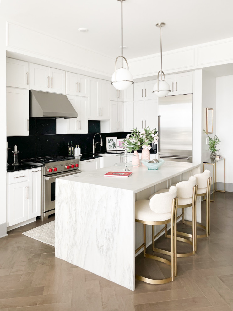 Example of a mid-sized transitional l-shaped medium tone wood floor and brown floor kitchen design in Nashville with a farmhouse sink, white cabinets, quartz countertops, black backsplash, stainless steel appliances and white countertops