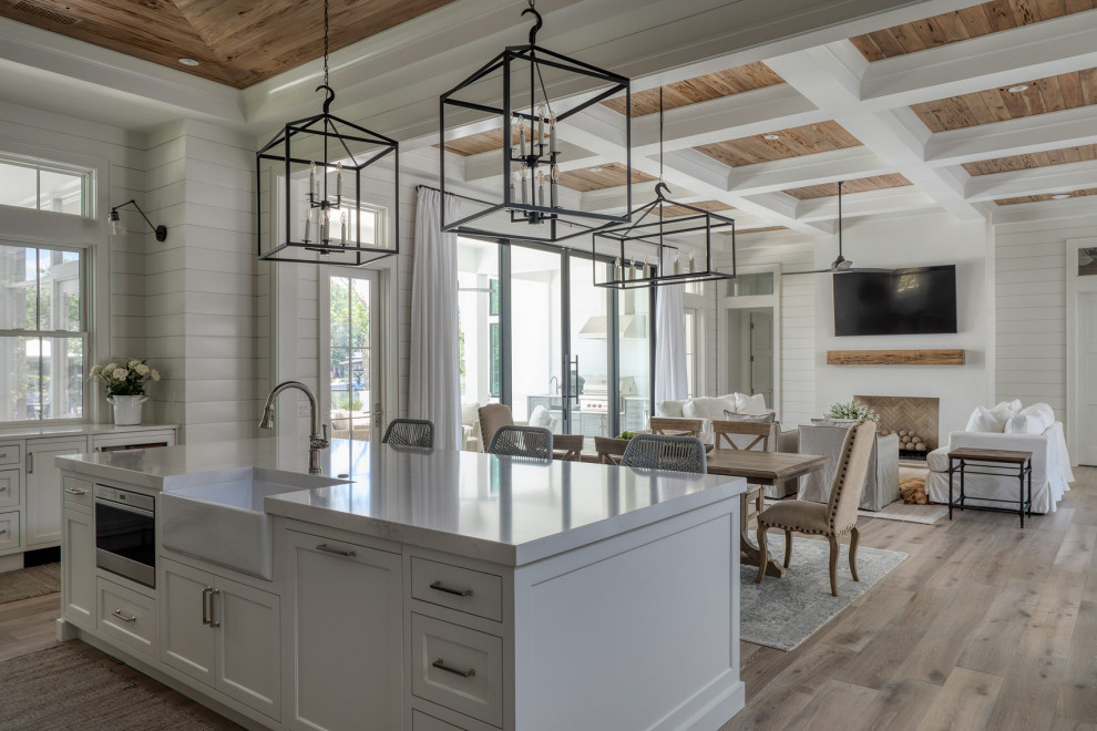 Example of a large transitional u-shaped light wood floor, beige floor and coffered ceiling open concept kitchen design with a farmhouse sink, recessed-panel cabinets, white cabinets, marble countertops, white backsplash, ceramic backsplash, white appliances, an island and white countertops