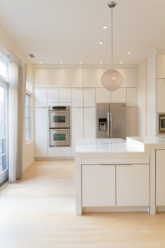 Photo of a large modern galley open plan kitchen in Chicago with a farmhouse sink, flat-panel cabinets, white cabinets, marble benchtops, white splashback, porcelain splashback, stainless steel appliances, light hardwood floors and with island.