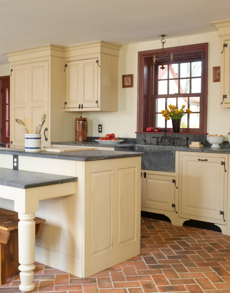 This is an example of a mid-sized country single-wall eat-in kitchen in Philadelphia with a farmhouse sink, raised-panel cabinets, with island, brick floors, panelled appliances, beige cabinets, concrete benchtops, red floor and grey benchtop.