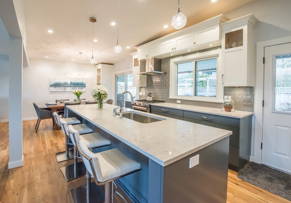 This is an example of a mid-sized contemporary l-shaped open plan kitchen in Vancouver with an undermount sink, shaker cabinets, white cabinets, quartzite benchtops, grey splashback, subway tile splashback, stainless steel appliances, medium hardwood floors, with island and brown floor.