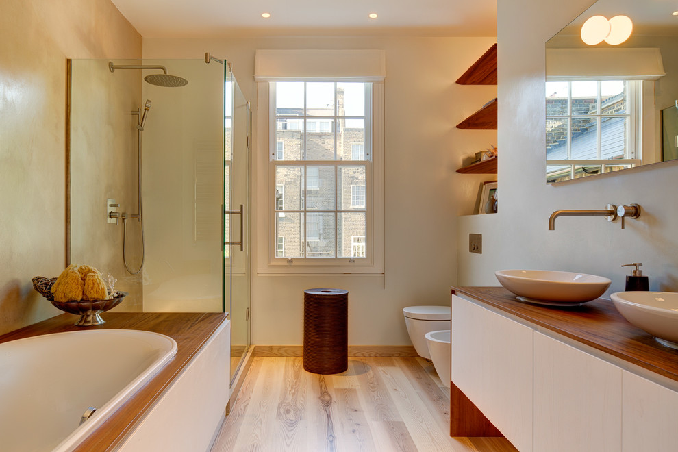 This is an example of a contemporary master bathroom in London with a vessel sink, flat-panel cabinets, white cabinets, wood benchtops, a drop-in tub, a corner shower, a bidet and light hardwood floors.
