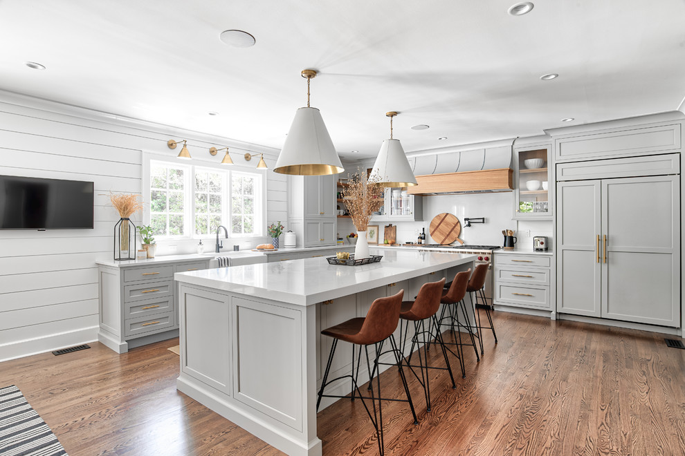 Photo of a country l-shaped kitchen in New York with a farmhouse sink, shaker cabinets, grey cabinets, white splashback, panelled appliances, medium hardwood floors, with island, white benchtop and quartz benchtops.
