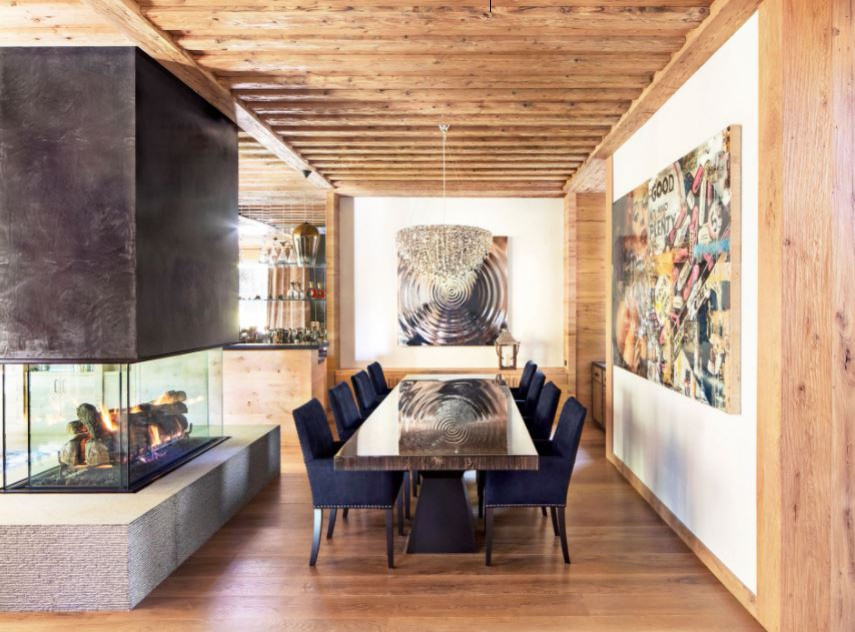 Cette photo montre une grande salle à manger ouverte sur le salon rétro avec un mur beige, parquet clair, une cheminée double-face, un manteau de cheminée en béton, un sol marron et poutres apparentes.
