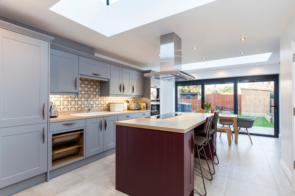 This is an example of a transitional galley kitchen in Surrey with a drop-in sink, shaker cabinets, blue cabinets, wood benchtops, multi-coloured splashback, stainless steel appliances and with island.