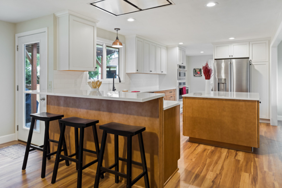 Mid-sized transitional l-shaped medium tone wood floor and brown floor eat-in kitchen photo with a farmhouse sink, shaker cabinets, white cabinets, quartz countertops, white backsplash, subway tile backsplash, stainless steel appliances, an island and white countertops