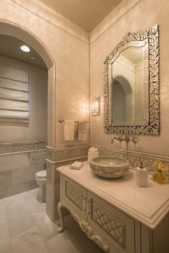 Photo of a mid-sized mediterranean powder room in Phoenix with a vessel sink, furniture-like cabinets, white cabinets, mosaic tile, beige walls, gray tile, a two-piece toilet, marble floors and solid surface benchtops.