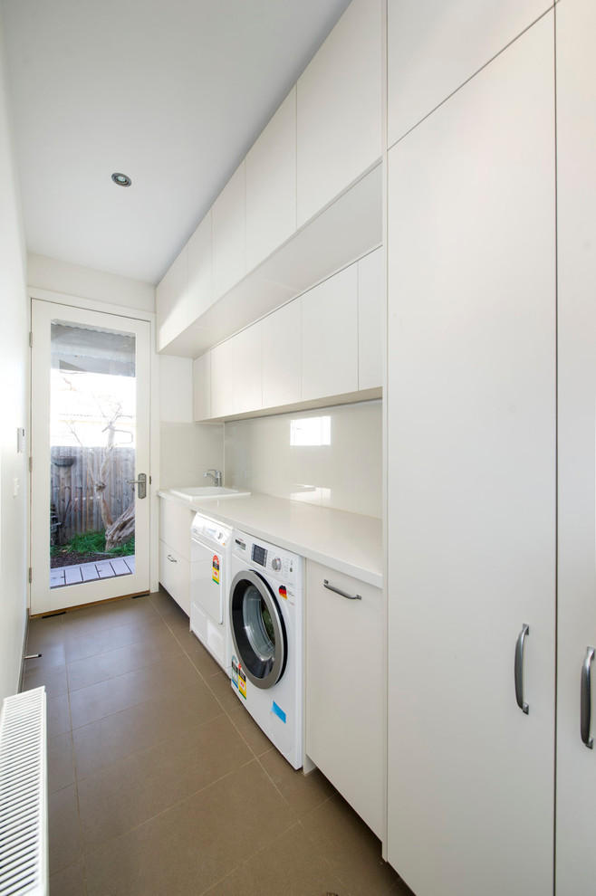 This is an example of a large contemporary galley dedicated laundry room in Melbourne with an integrated sink, white cabinets, quartz benchtops, white walls, ceramic floors, a side-by-side washer and dryer, brown floor and white benchtop.