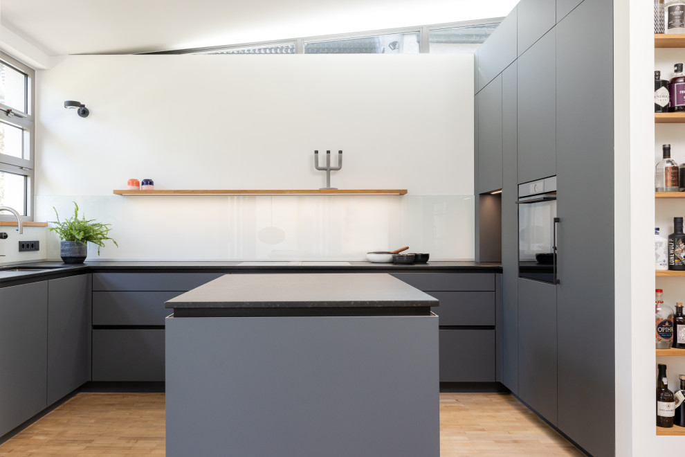 Inspiration for an expansive contemporary u-shaped open plan kitchen in Frankfurt with an undermount sink, flat-panel cabinets, grey cabinets, granite benchtops, white splashback, glass sheet splashback, black appliances, medium hardwood floors, with island, brown floor, black benchtop and recessed.