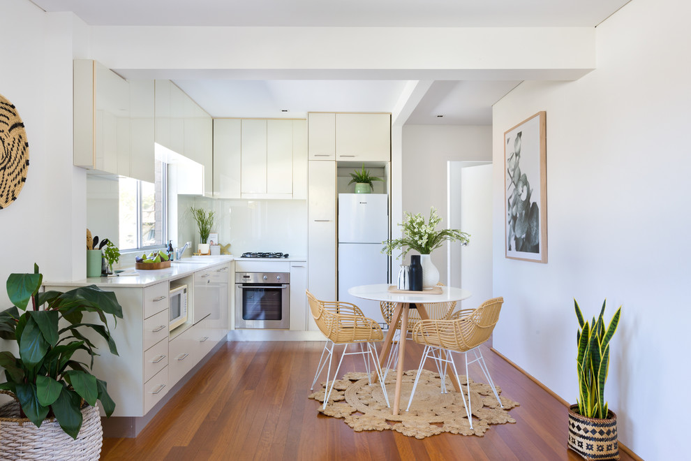 Photo of a contemporary l-shaped eat-in kitchen in Sydney with an undermount sink, flat-panel cabinets, white cabinets, white splashback, white appliances, medium hardwood floors, no island, brown floor and white benchtop.