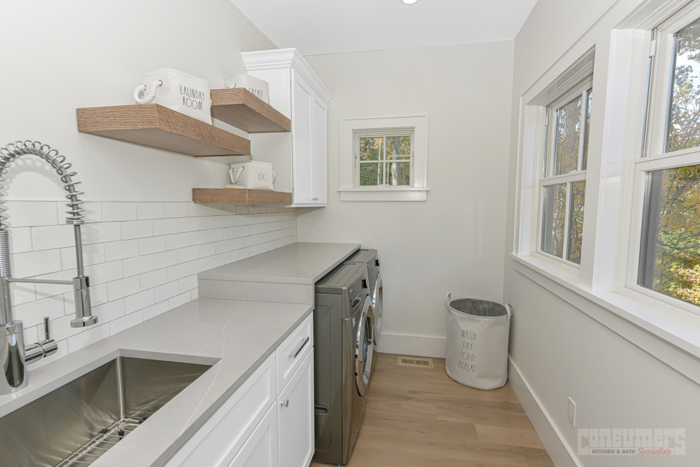 Small midcentury single-wall dedicated laundry room in New York with a drop-in sink, recessed-panel cabinets, white cabinets, quartz benchtops, white splashback, subway tile splashback, white walls, medium hardwood floors, a side-by-side washer and dryer, brown floor and grey benchtop.
