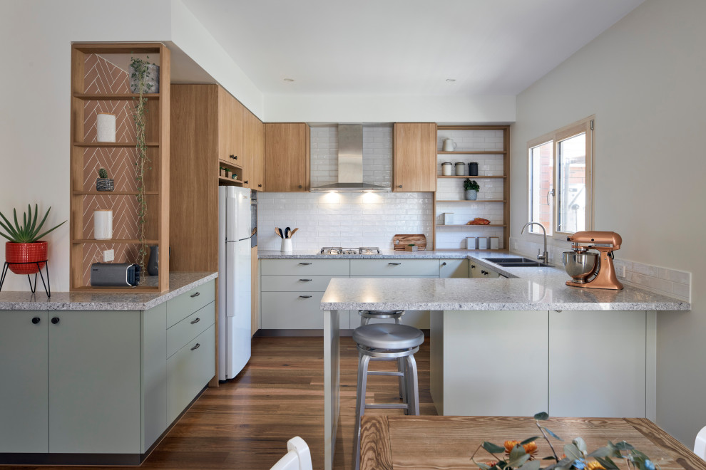 This is an example of a contemporary u-shaped eat-in kitchen in Melbourne with a double-bowl sink, flat-panel cabinets, grey cabinets, white splashback, subway tile splashback, white appliances, medium hardwood floors, with island, brown floor and multi-coloured benchtop.