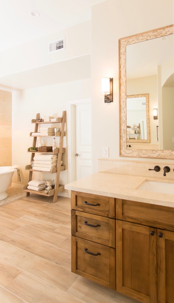 Photo of a mid-sized mediterranean master bathroom in Phoenix with shaker cabinets, medium wood cabinets, a freestanding tub, a one-piece toilet, beige tile, travertine, beige walls, porcelain floors, an undermount sink, marble benchtops, beige floor and a hinged shower door.