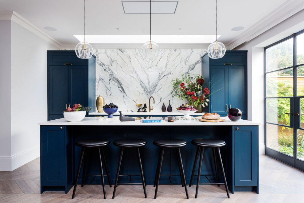 Large contemporary kitchen in London with shaker cabinets, blue cabinets, quartzite benchtops, white splashback, marble splashback, black appliances, with island, brown floor and white benchtop.