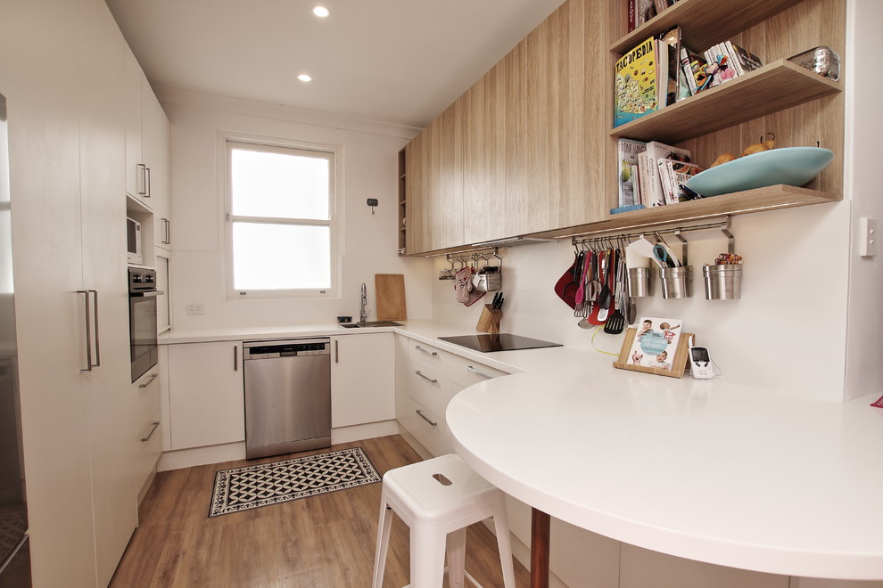 Photo of a mid-sized beach style u-shaped eat-in kitchen in Sydney with a single-bowl sink, flat-panel cabinets, white cabinets, solid surface benchtops, white splashback, ceramic splashback, stainless steel appliances, vinyl floors, a peninsula and brown floor.