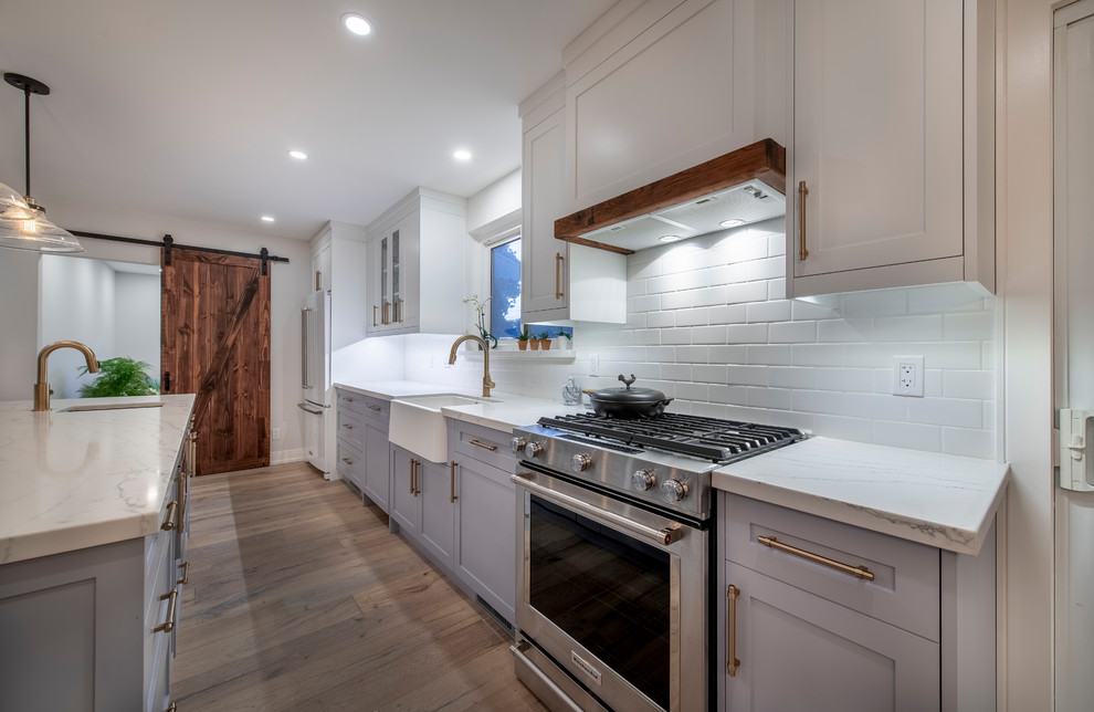This is an example of a contemporary galley eat-in kitchen in Toronto with a farmhouse sink, shaker cabinets, white cabinets, quartz benchtops, white splashback, brick splashback, stainless steel appliances, painted wood floors, with island, brown floor and white benchtop.