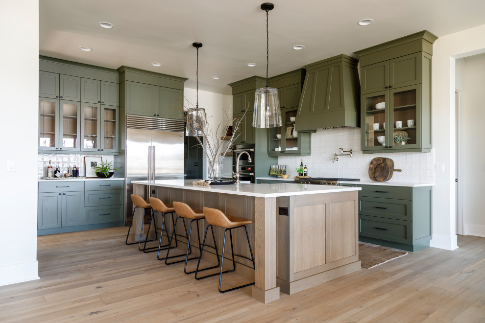 Photo of a mid-sized country l-shaped eat-in kitchen in Boise with a farmhouse sink, shaker cabinets, green cabinets, quartz benchtops, white splashback, ceramic splashback, stainless steel appliances, medium hardwood floors, with island and white benchtop.