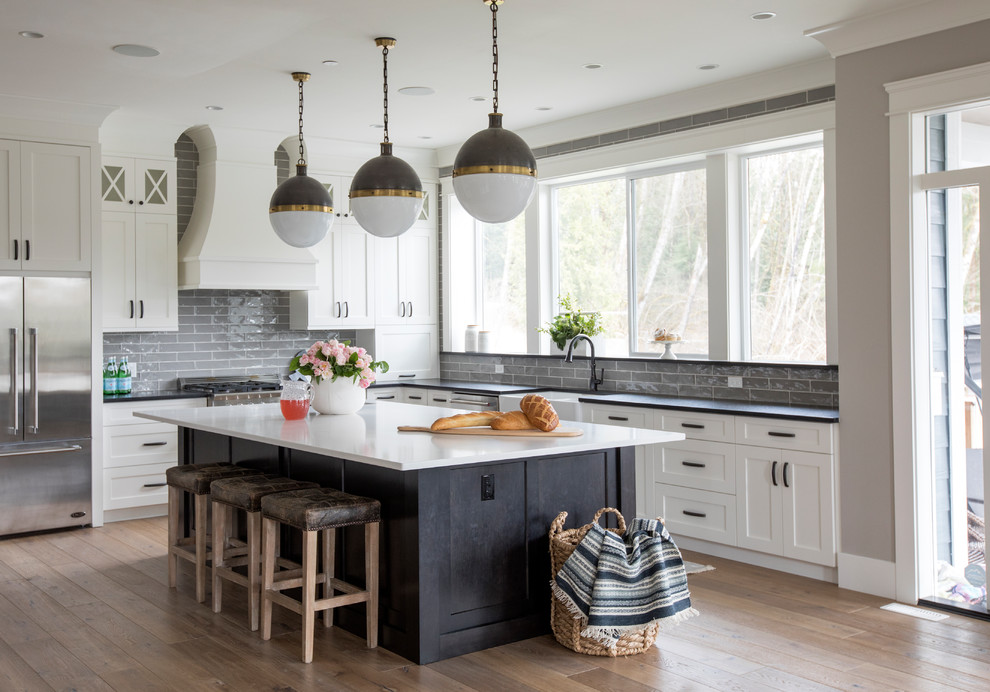 This is an example of a country u-shaped open plan kitchen in Other with a farmhouse sink, shaker cabinets, white cabinets, solid surface benchtops, grey splashback, stainless steel appliances, light hardwood floors, with island, brown floor and glass tile splashback.