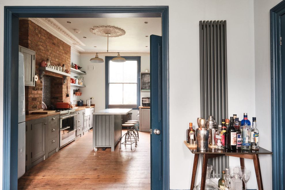 Large eclectic galley open plan kitchen in London with a farmhouse sink, shaker cabinets, grey cabinets, zinc benchtops, blue splashback, ceramic splashback, coloured appliances, medium hardwood floors, with island and grey benchtop.