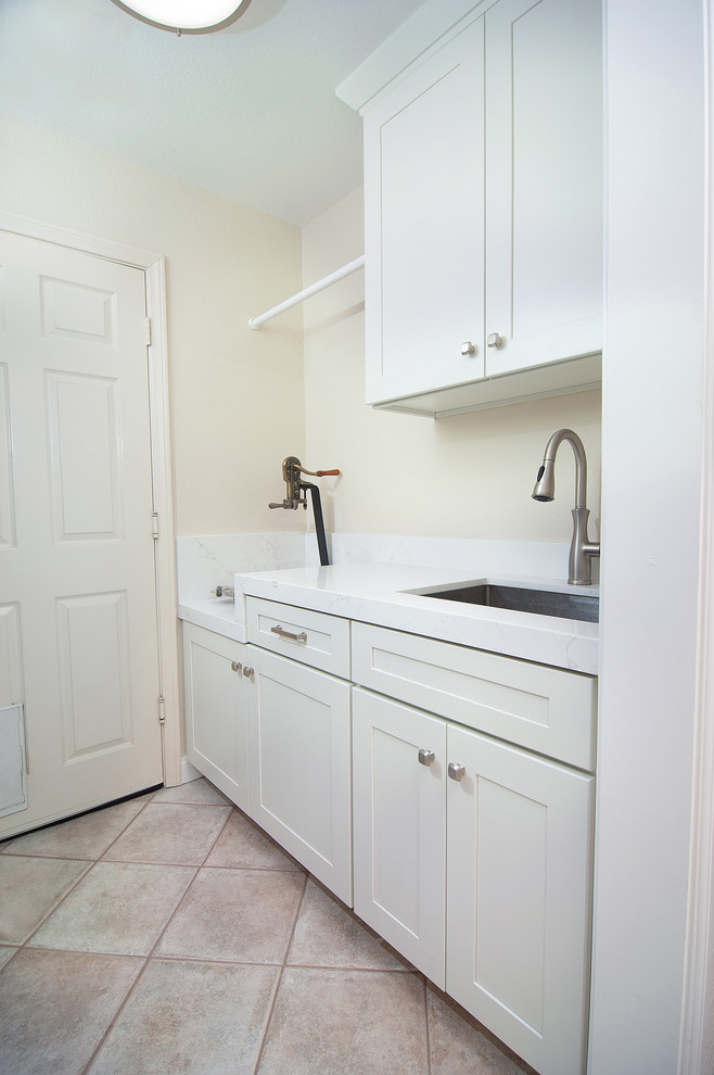 Photo of a small transitional galley dedicated laundry room in San Diego with an undermount sink, shaker cabinets, white cabinets, quartzite benchtops, beige walls, travertine floors, a side-by-side washer and dryer, brown floor and white benchtop.