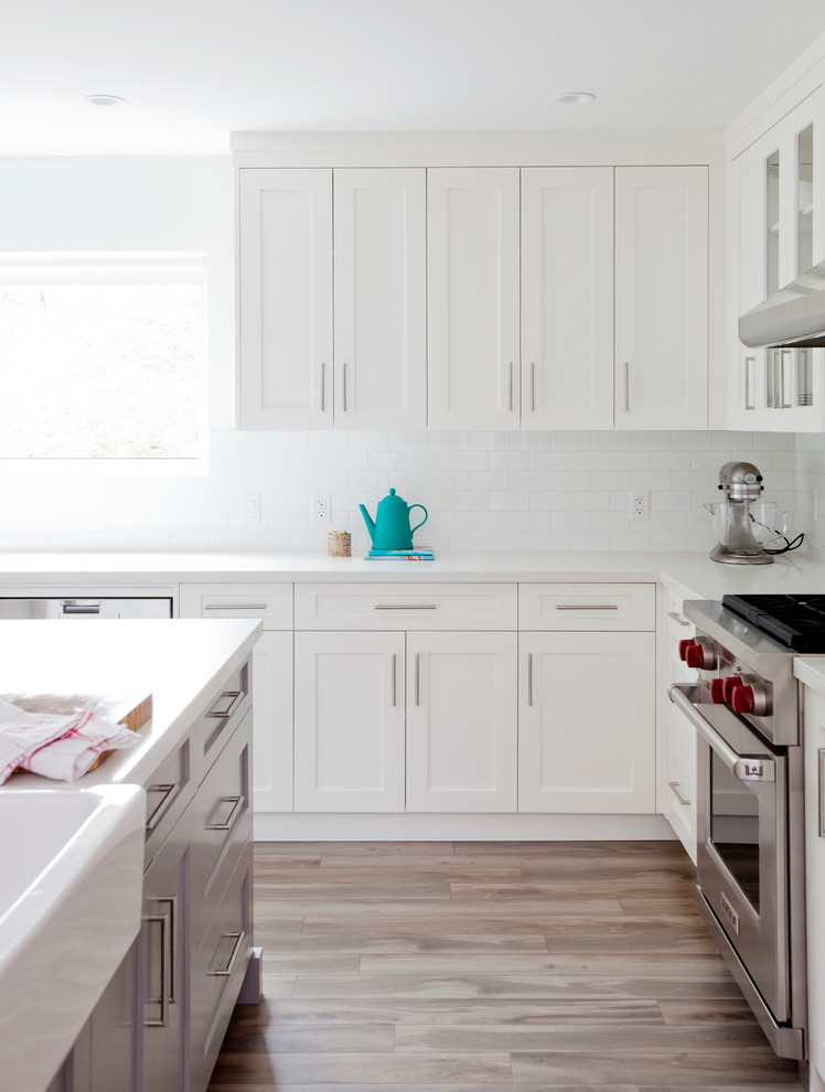 Inspiration for a large transitional l-shaped eat-in kitchen in Vancouver with a farmhouse sink, shaker cabinets, white cabinets, solid surface benchtops, white splashback, subway tile splashback, stainless steel appliances, light hardwood floors and with island.