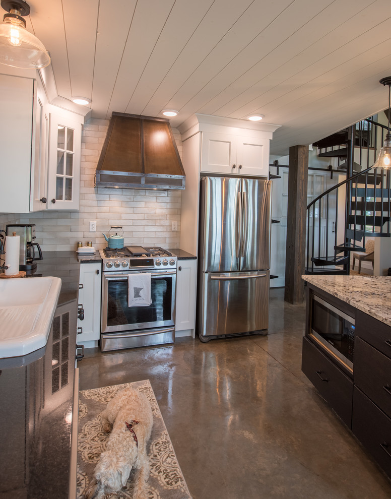 Example of a mid-sized mountain style l-shaped concrete floor and gray floor open concept kitchen design in Minneapolis with a drop-in sink, shaker cabinets, white cabinets, solid surface countertops, beige backsplash, subway tile backsplash, stainless steel appliances, an island and black countertops