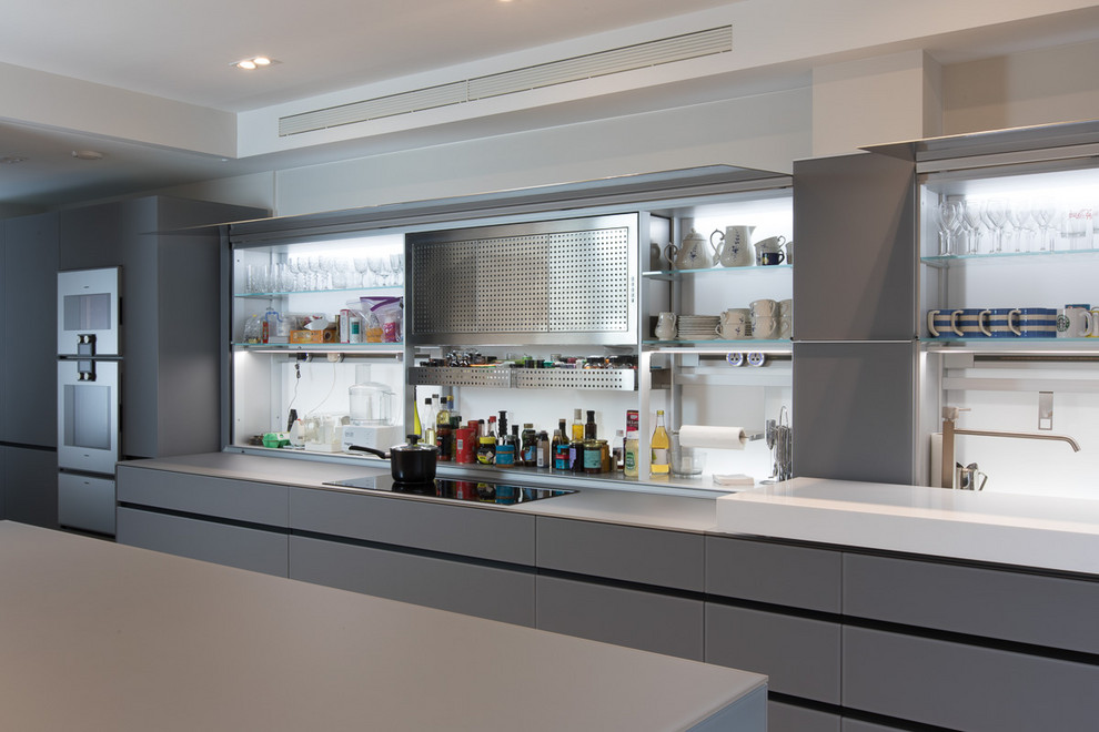 This is an example of a mid-sized modern galley eat-in kitchen in London with a drop-in sink, glass-front cabinets, grey cabinets, glass benchtops, white splashback, glass sheet splashback, light hardwood floors, with island and grey benchtop.