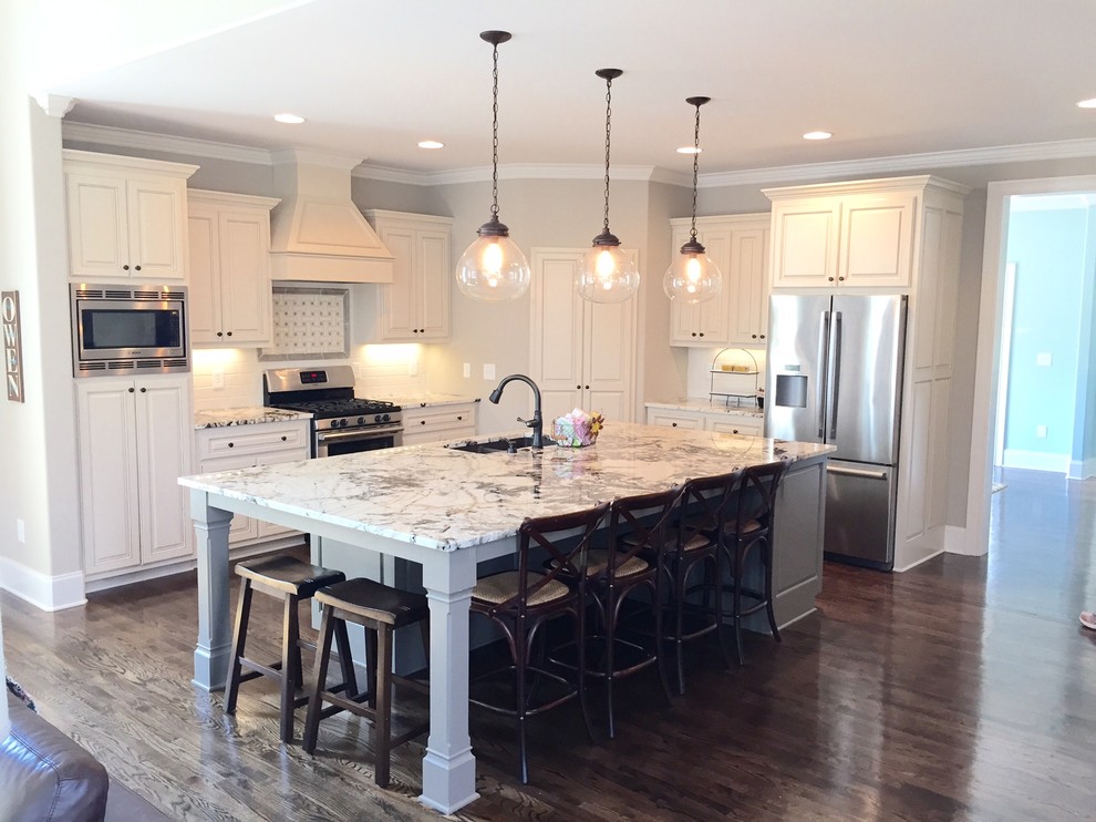 Example of a large transitional medium tone wood floor and brown floor open concept kitchen design with an undermount sink, raised-panel cabinets, white cabinets, granite countertops, white backsplash, subway tile backsplash, stainless steel appliances and an island