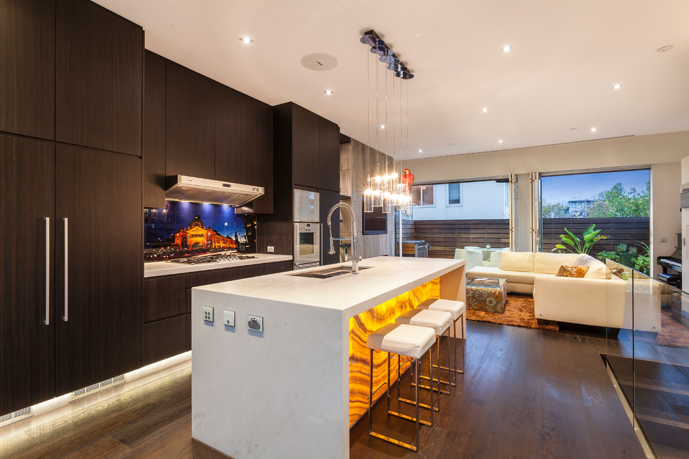 This is an example of a mid-sized contemporary single-wall open plan kitchen in Melbourne with flat-panel cabinets, dark wood cabinets, dark hardwood floors, with island, brown floor, a double-bowl sink, marble benchtops, multi-coloured splashback, glass sheet splashback, white appliances and white benchtop.