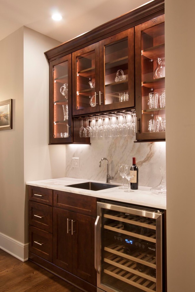 This is an example of a large transitional single-wall wet bar in Other with glass-front cabinets, medium wood cabinets, marble benchtops, beige splashback, marble splashback, medium hardwood floors, brown floor and an undermount sink.