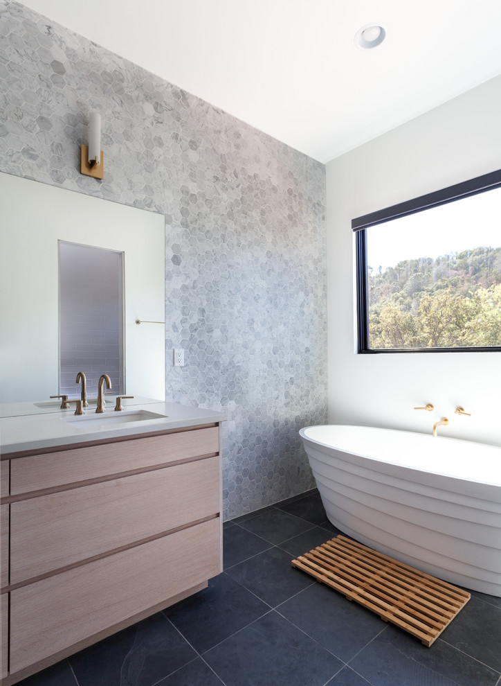 This is an example of a contemporary bathroom in Sacramento with flat-panel cabinets, light wood cabinets, a freestanding tub, gray tile, stone tile, white walls and an undermount sink.