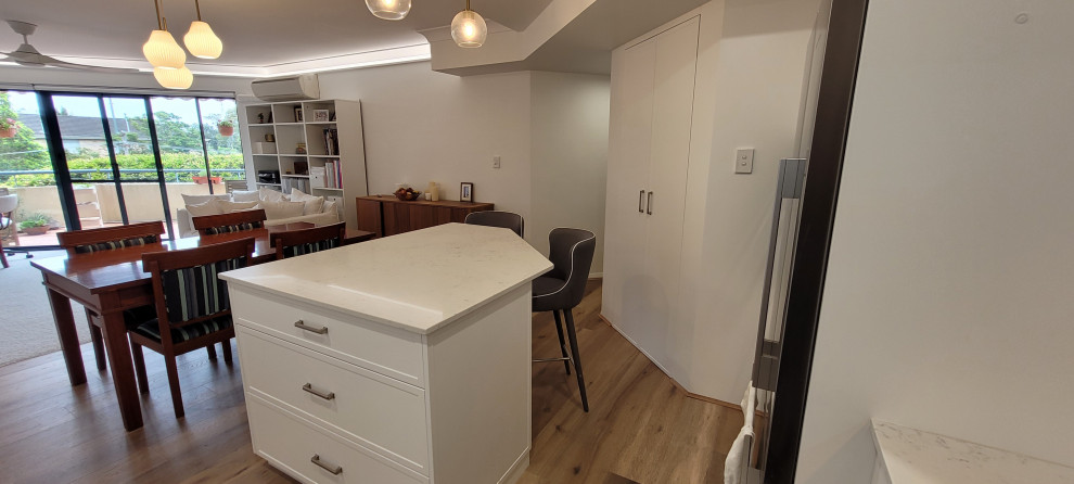 Mid-sized contemporary l-shaped eat-in kitchen in Sydney with a single-bowl sink, shaker cabinets, white cabinets, quartz benchtops, white splashback, engineered quartz splashback, stainless steel appliances, laminate floors, with island, brown floor, white benchtop and recessed.