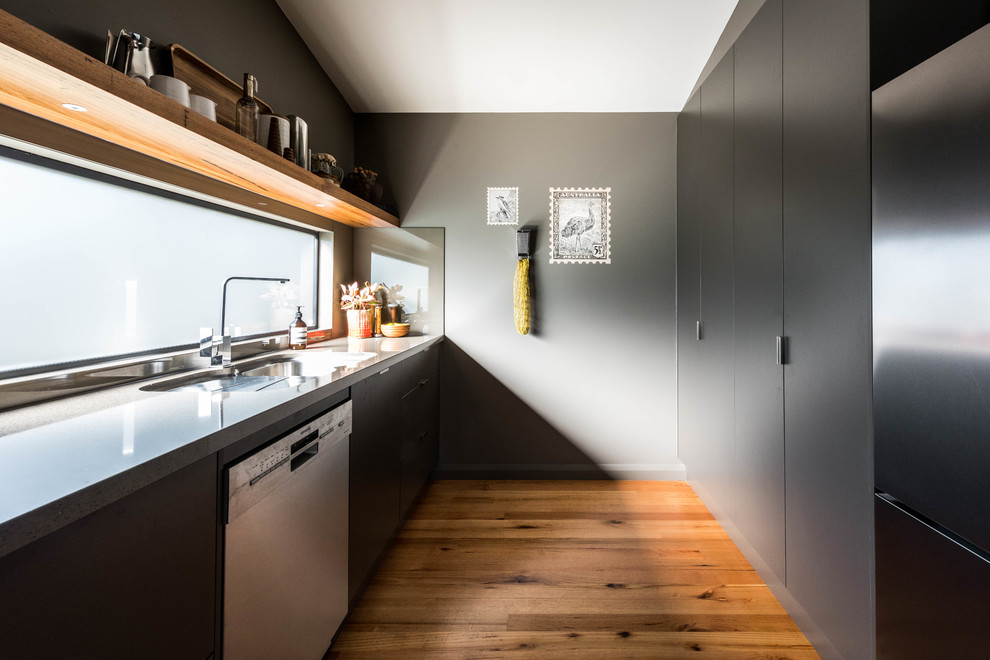 Inspiration for a contemporary galley separate kitchen in Melbourne with a drop-in sink, flat-panel cabinets, black cabinets, window splashback, stainless steel appliances, medium hardwood floors, no island, brown floor and grey benchtop.