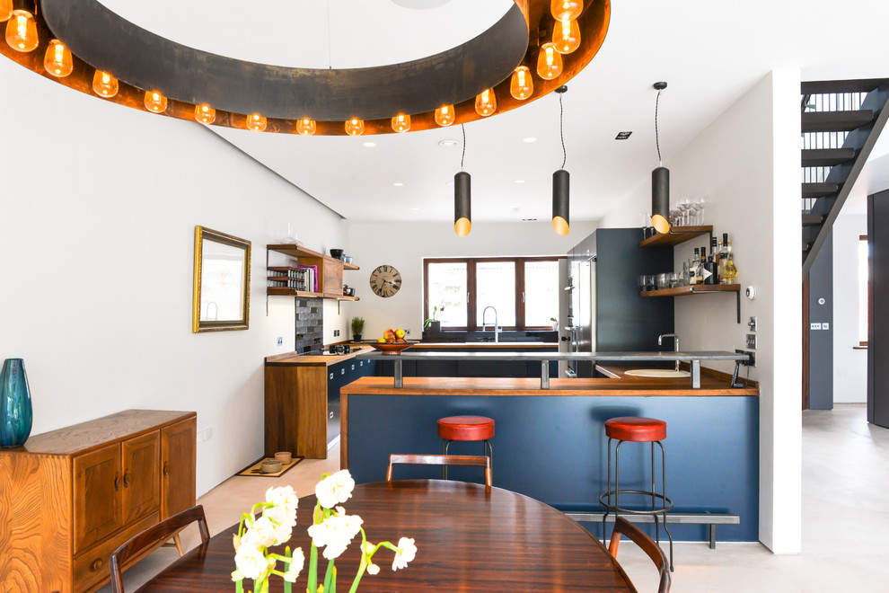 Photo of a mid-sized contemporary open plan kitchen in Sussex with a drop-in sink, flat-panel cabinets, blue cabinets, wood benchtops, black splashback, ceramic splashback, black appliances, concrete floors and with island.