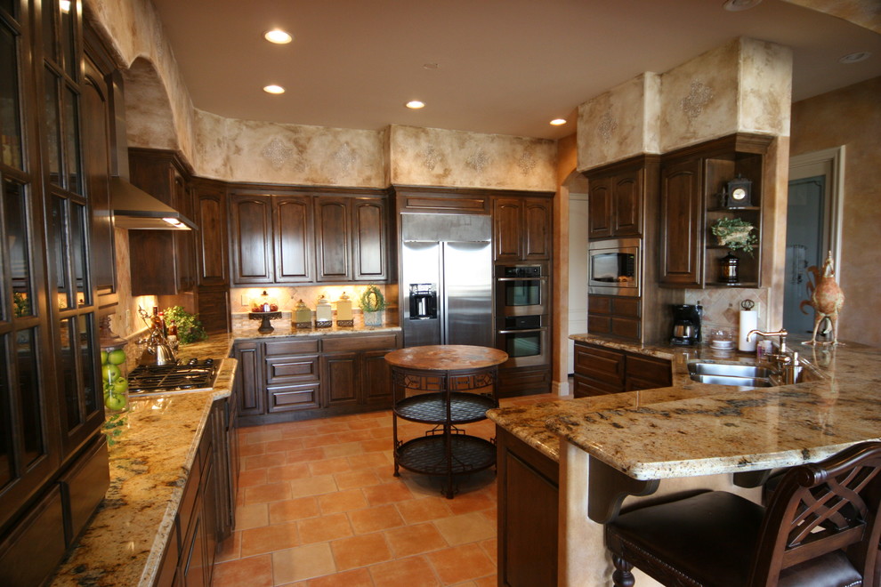 This is an example of a mediterranean l-shaped eat-in kitchen in Other with a double-bowl sink, raised-panel cabinets, dark wood cabinets, granite benchtops, orange splashback, terra-cotta splashback, stainless steel appliances, terra-cotta floors, a peninsula, orange floor and multi-coloured benchtop.