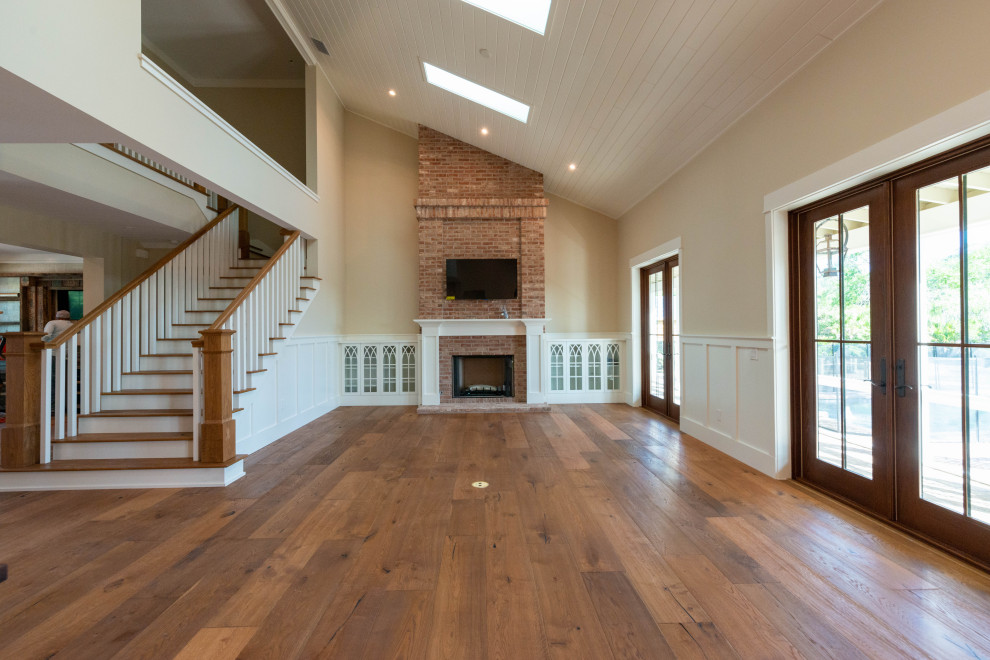 Example of a large farmhouse formal and open concept medium tone wood floor, brown floor, vaulted ceiling and wainscoting living room design in Miami with beige walls, a standard fireplace, a brick fireplace and a wall-mounted tv