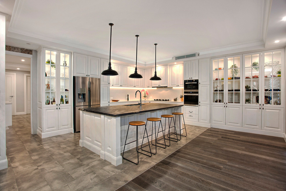 Large contemporary l-shaped kitchen pantry in Sydney with a double-bowl sink, beaded inset cabinets, white cabinets, white splashback, stainless steel appliances, with island and black benchtop.