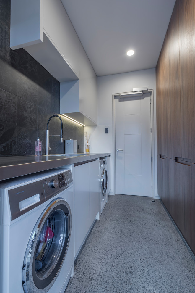 This is an example of a large modern galley dedicated laundry room in Auckland with an integrated sink, flat-panel cabinets, dark wood cabinets, stainless steel benchtops, grey walls, concrete floors, a side-by-side washer and dryer and grey floor.