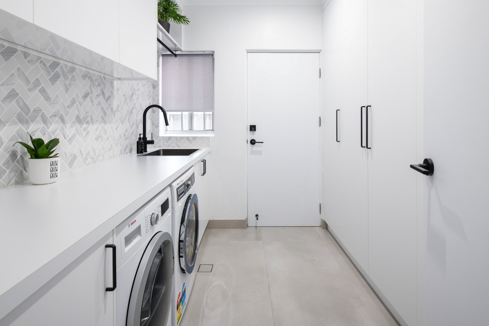 This is an example of a large modern galley dedicated laundry room in Sydney with a drop-in sink, flat-panel cabinets, white cabinets, laminate benchtops, grey splashback, mosaic tile splashback, white walls, ceramic floors, a side-by-side washer and dryer, grey floor and white benchtop.