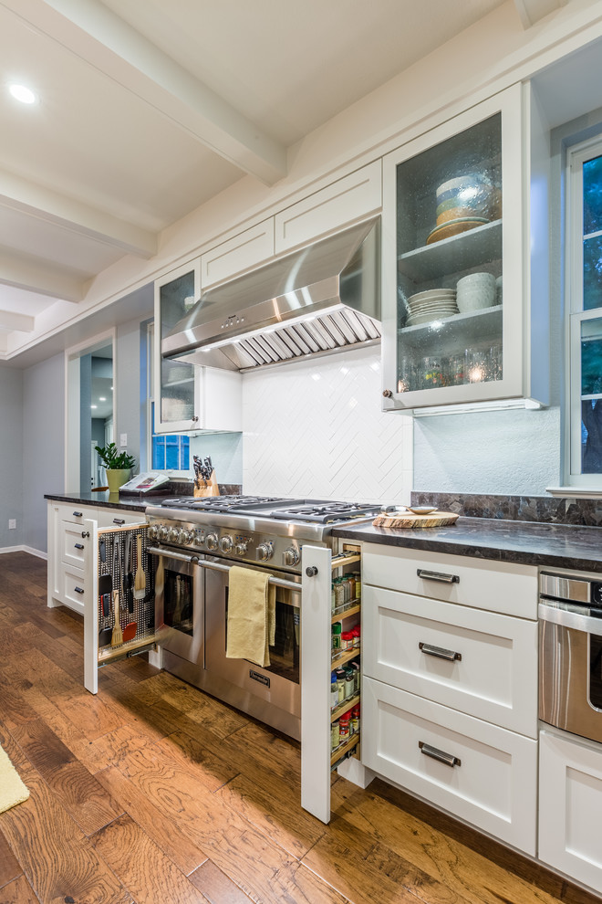 Example of a mid-sized transitional l-shaped medium tone wood floor eat-in kitchen design in Austin with an undermount sink, shaker cabinets, white cabinets, granite countertops, white backsplash, ceramic backsplash, stainless steel appliances and an island