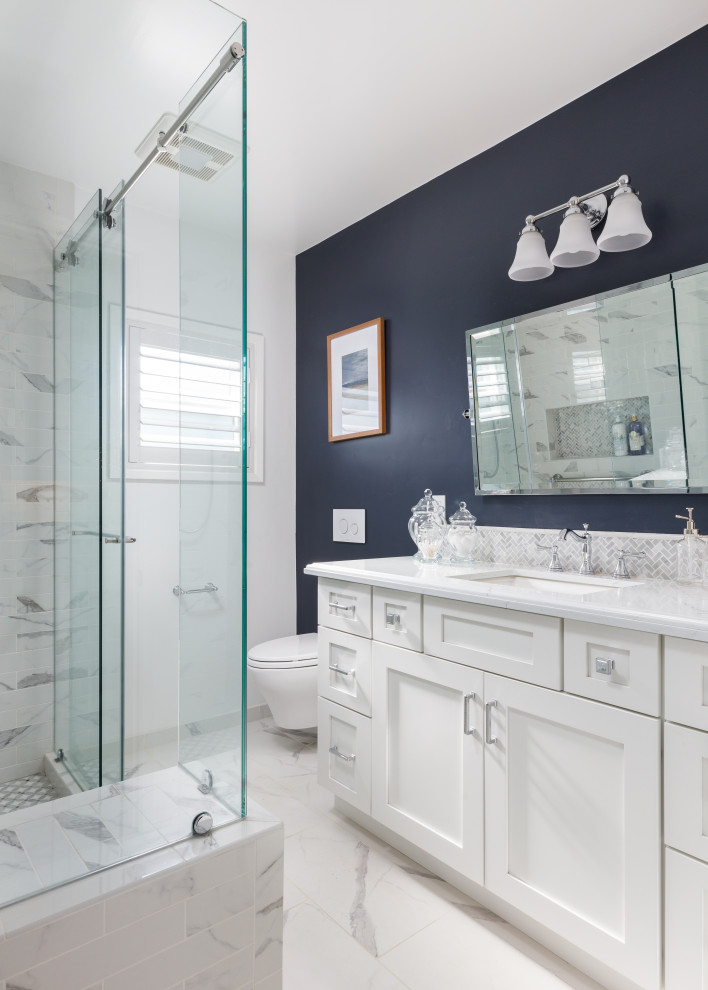 This is an example of a mid-sized country master bathroom in Los Angeles with recessed-panel cabinets, white cabinets, an alcove shower, a wall-mount toilet, gray tile, porcelain tile, blue walls, porcelain floors, an undermount sink, engineered quartz benchtops, white floor, a sliding shower screen, white benchtops, a shower seat, a single vanity and a built-in vanity.