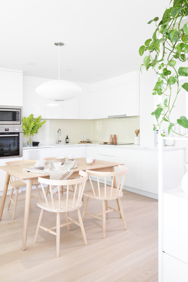 This is an example of a scandinavian l-shaped eat-in kitchen in Vancouver with flat-panel cabinets, white cabinets, beige splashback, glass sheet splashback, stainless steel appliances, light hardwood floors, no island, beige floor and white benchtop.