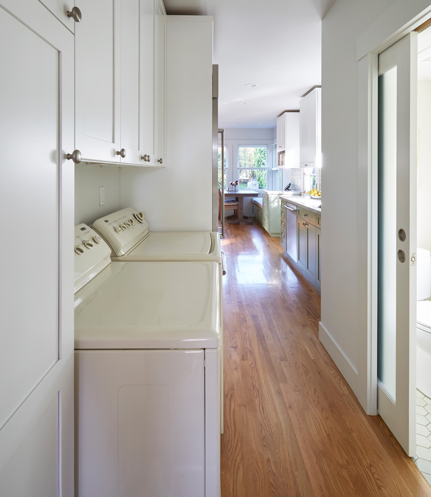 Mid-sized transitional galley laundry room in San Francisco with shaker cabinets, grey splashback, subway tile splashback, light hardwood floors, brown floor, white cabinets and a side-by-side washer and dryer.