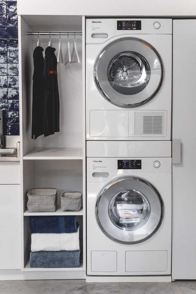 Photo of a large modern single-wall dedicated laundry room in Other with an integrated sink, white cabinets, stainless steel benchtops, blue splashback, ceramic splashback, blue walls, ceramic floors, a stacked washer and dryer, grey floor and grey benchtop.