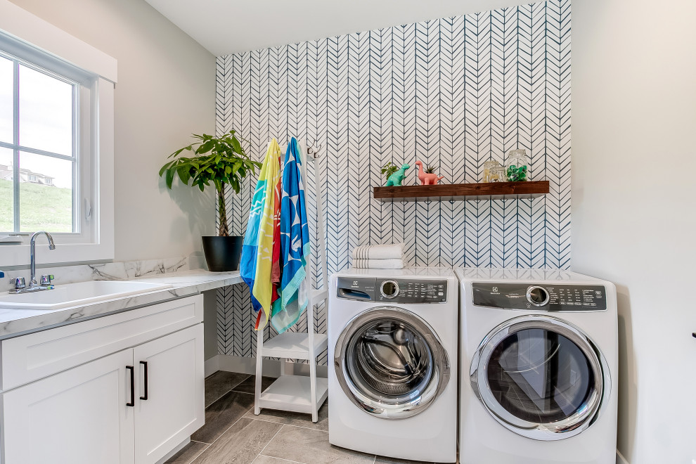 Mid-sized transitional l-shaped dedicated laundry room in Other with a drop-in sink, shaker cabinets, white cabinets, grey walls, porcelain floors, a side-by-side washer and dryer, grey floor, grey benchtop and wallpaper.