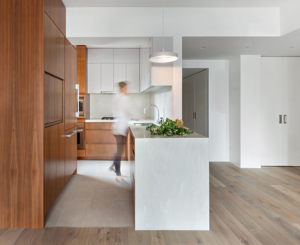 Photo of a small contemporary l-shaped open plan kitchen in New York with an undermount sink, flat-panel cabinets, medium wood cabinets, solid surface benchtops, stainless steel appliances, porcelain floors, a peninsula, beige floor, white splashback and white benchtop.