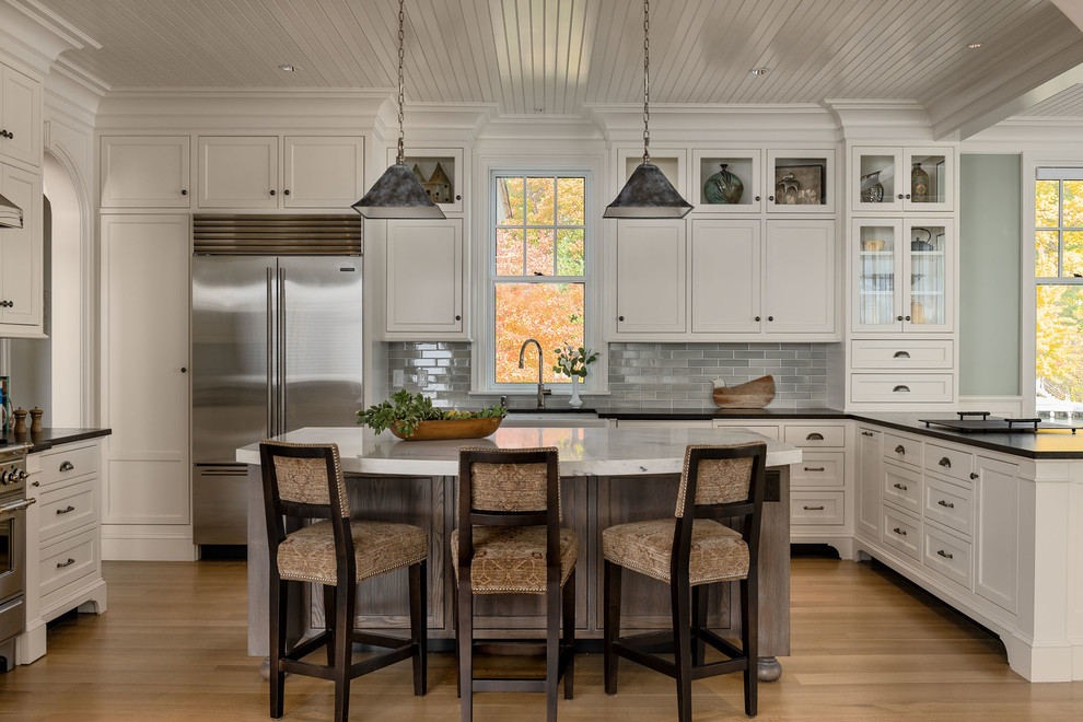 Beach style kitchen in Boston with a farmhouse sink, shaker cabinets, white cabinets, grey splashback, subway tile splashback, stainless steel appliances, light hardwood floors, with island, beige floor and black benchtop.