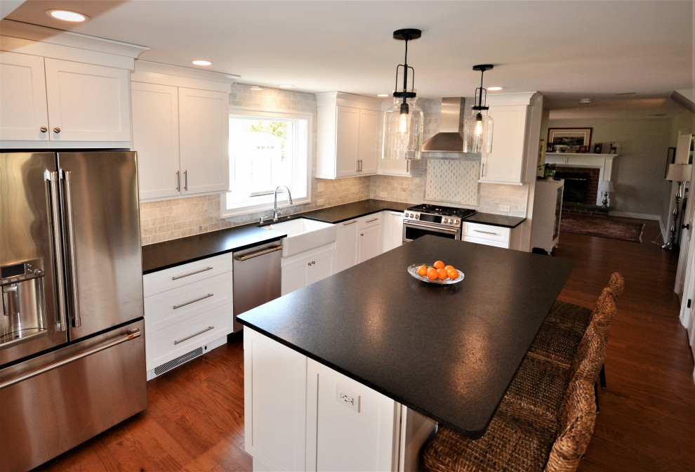 This is an example of a large transitional single-wall eat-in kitchen in Philadelphia with a farmhouse sink, recessed-panel cabinets, white cabinets, granite benchtops, grey splashback, brick splashback, stainless steel appliances, dark hardwood floors, with island, brown floor and black benchtop.